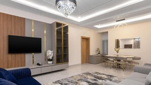 A modern living room with a flat-screen TV, a large bookshelf, a chandelier, and a dining area with a table and chairs. - STP Apartments (Abuja)