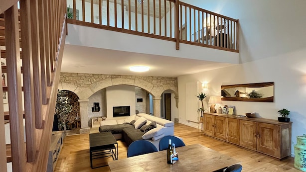 Un vaste espace de vie agrémenté d’un escalier en bois, d’une arche en pierre, d’une table à manger avec des chaises, et d’un coin cuisine doté de meubles en bois.