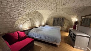 Une pièce chaleureuse avec un plafond en pierre, un lit recouvert de linge de maison blanc, un canapé rouge et un petit meuble.