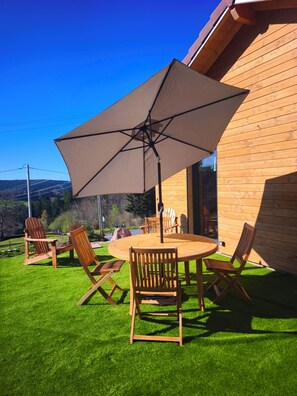 Eine Holzterrasse mit einem runden Tisch, Stühlen und einem Sonnenschirm, mit Blick auf ein bewaldetes Gebiet.