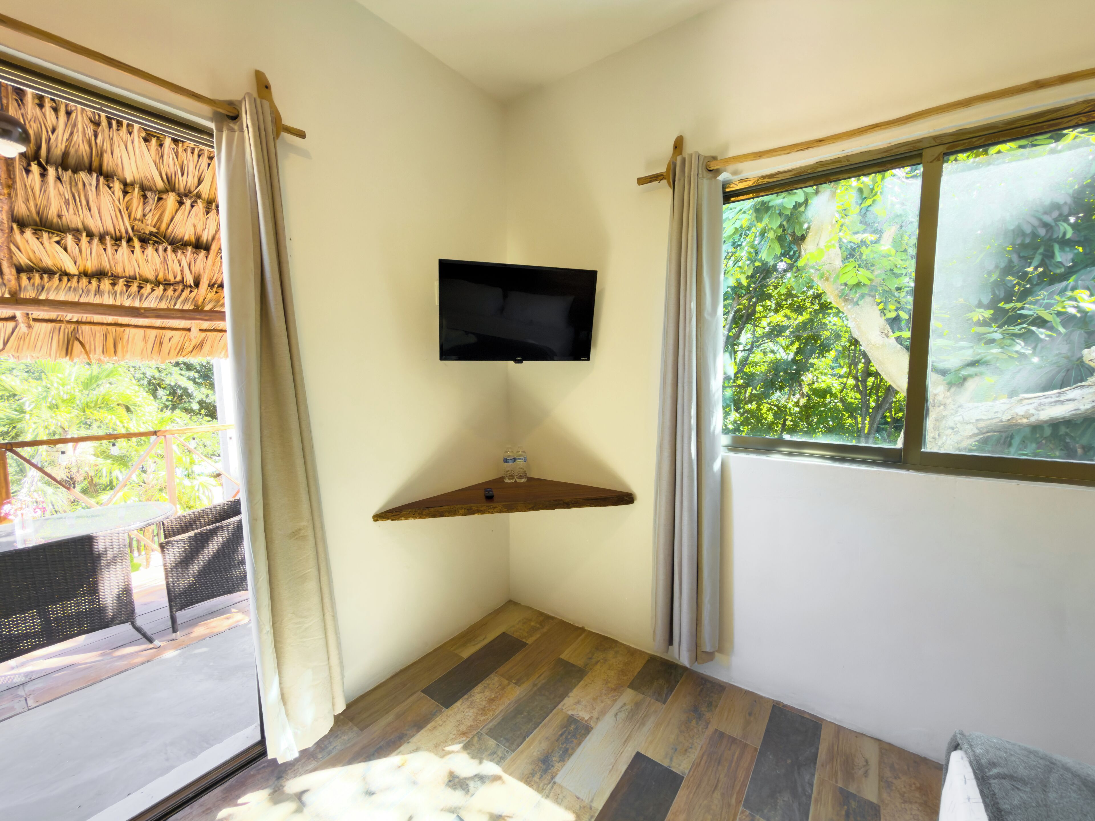 Ein Zimmer mit an der Wand montiertem Fernseher, einem Holzregal, einem Fenster mit Vorhängen und Blick ins Grüne.