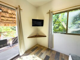 Una habitación con televisión montada en la pared, una estantería de madera, una ventana con cortinas y vistas a zonas verdes.