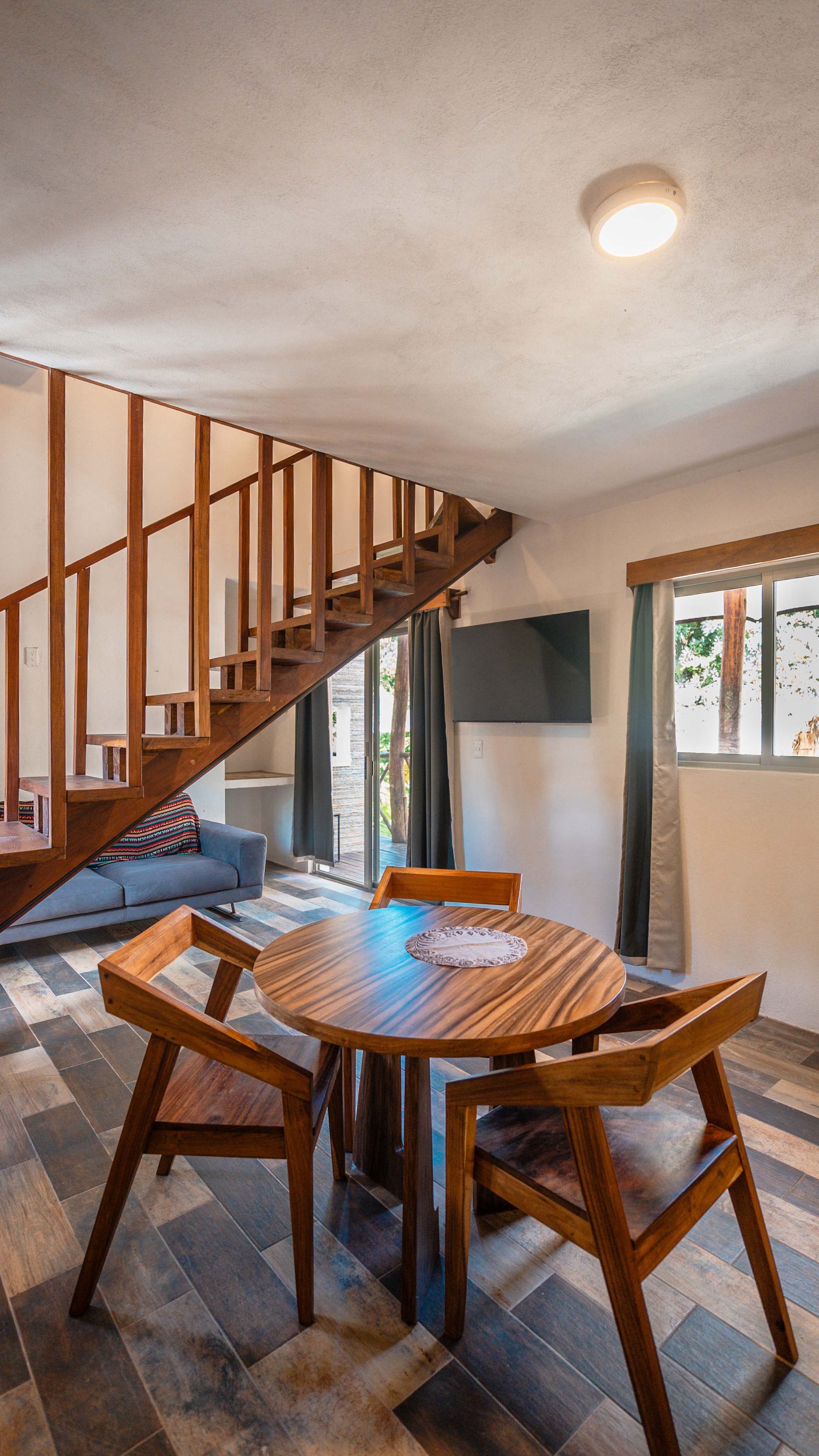 A dining area with a round wooden table and chairs, a staircase with wooden railings, and a view of the outdoors through a window.