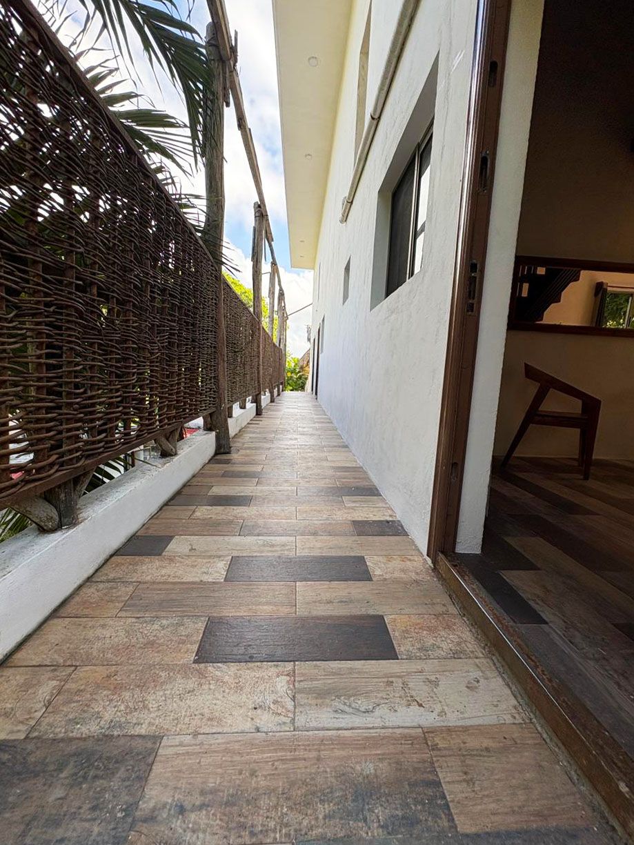 A narrow corridor with a patterned floor, a wicker fence on one side, and a building entrance on the other.