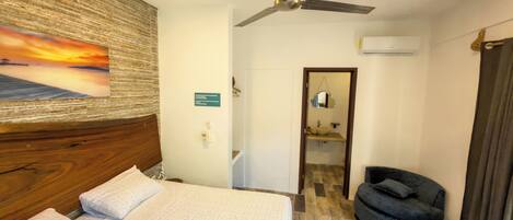 Habitación de hotel con cama, ventilador de techo, baño con lavamanos y espejo, y una silla.