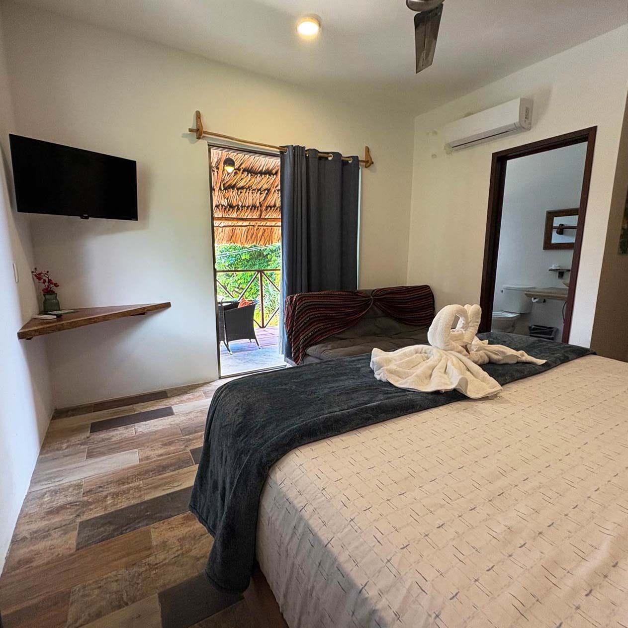 A hotel room with a bed, a TV, a small shelf, a door leading to a balcony, and a bathroom.