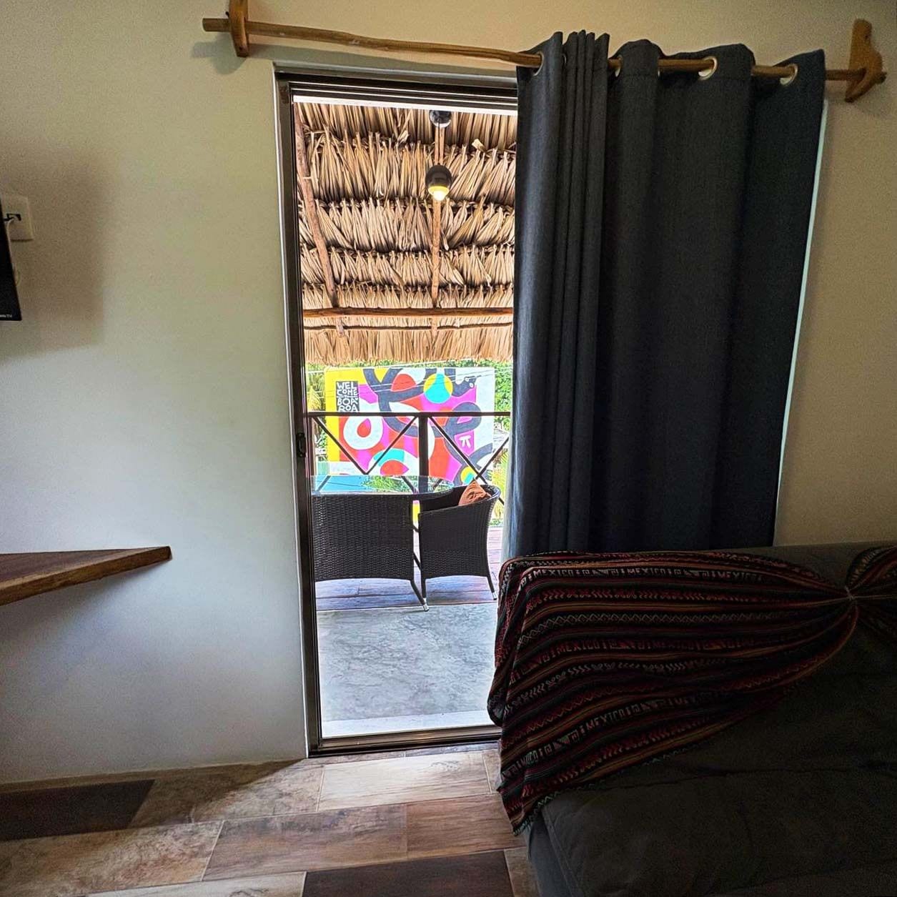 Une pièce comprenant un canapé, une étagère en bois, un plafond en chaume et donnant sur un espace extérieur aux couleurs vives.