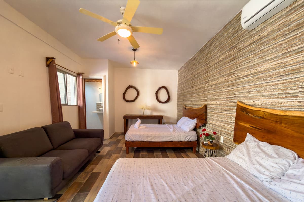 A bedroom with a bed, a wooden headboard, a nightstand, a small table with a vase of flowers, a ceiling fan, and a wall-mounted air conditioner.