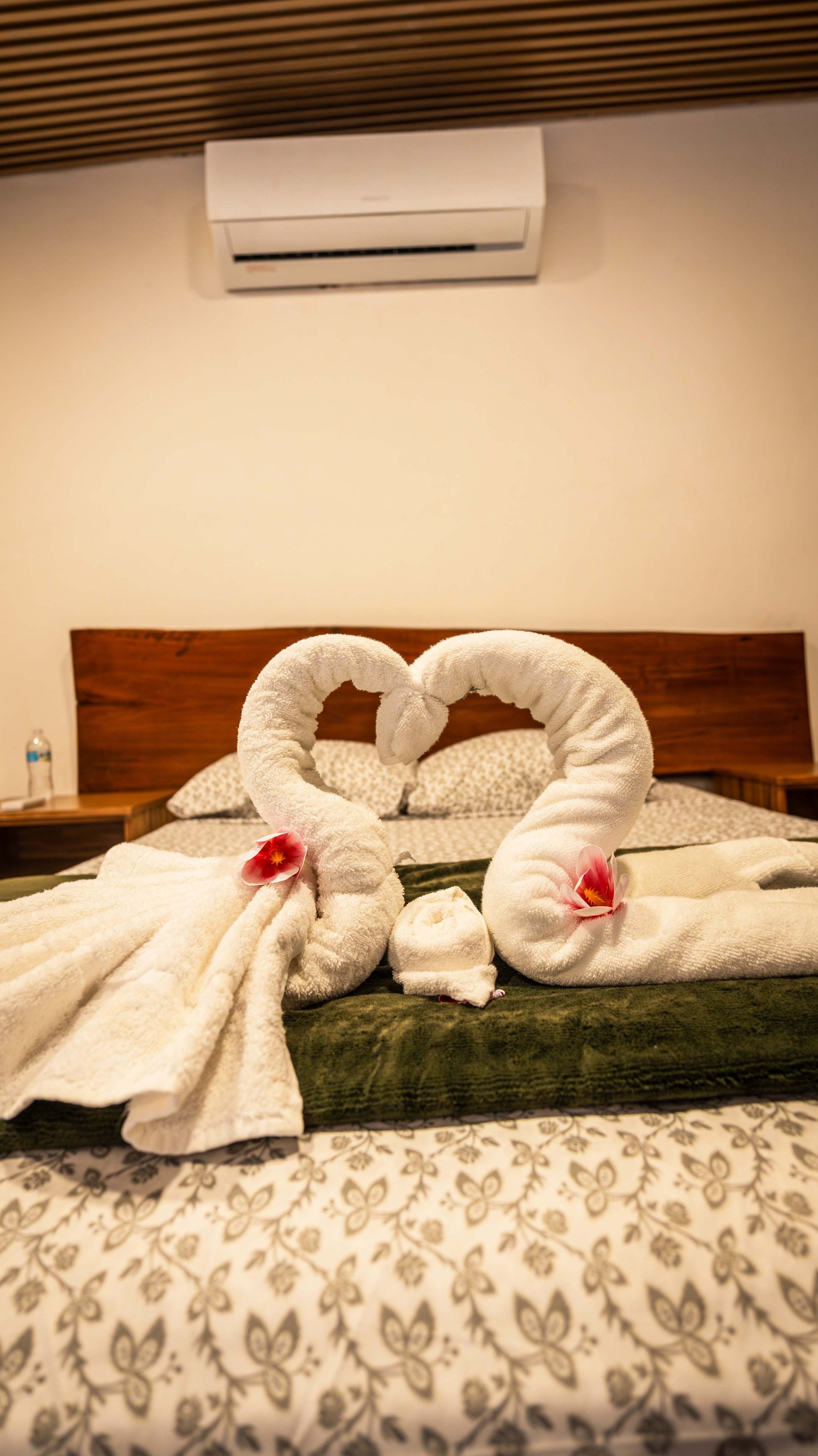 A bed with a heart-shaped towel arrangement, a wooden headboard, a patterned bedspread, and an air conditioning unit above.