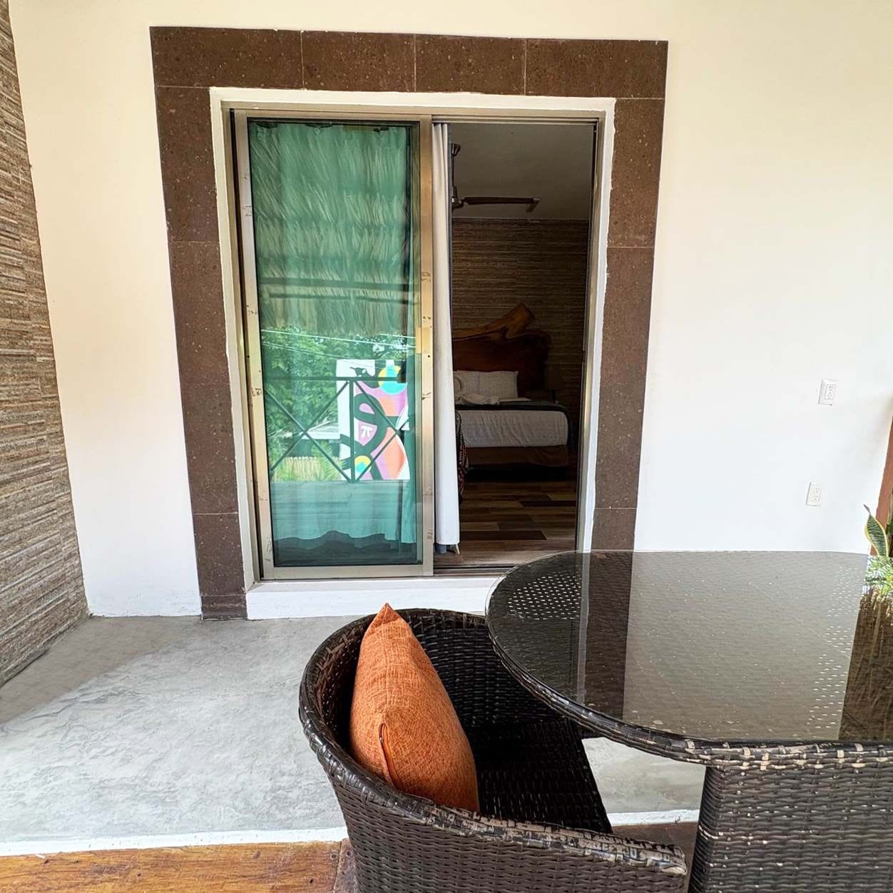 A room with a bed, a window with a green curtain, and a wicker chair with a cushion.