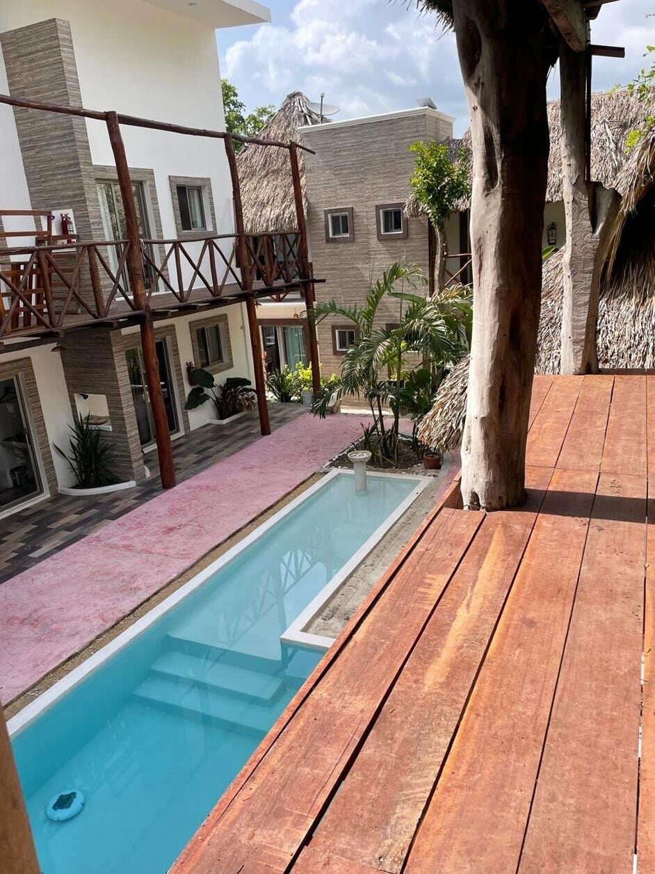 A pool area with a wooden deck, a thatched roof structure, and a building with multiple windows.