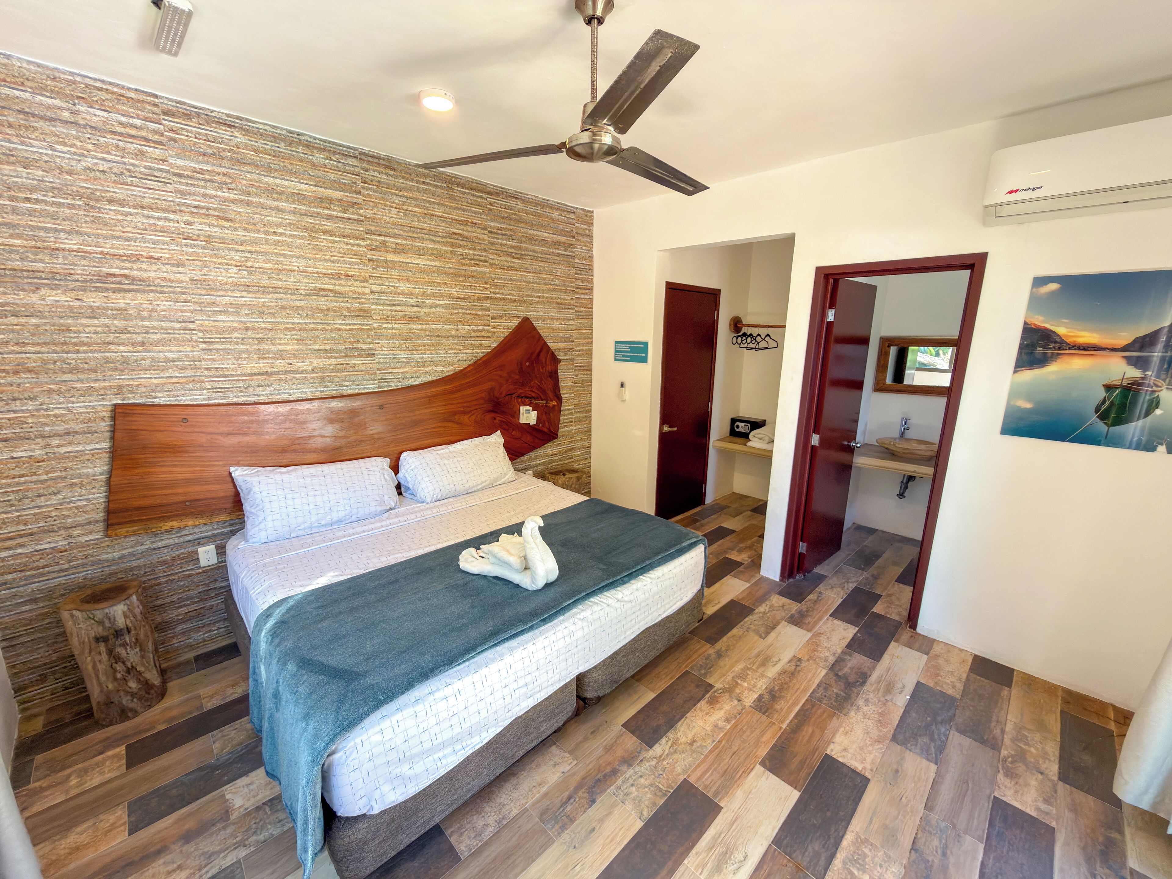 A bedroom with a wooden headboard, a bed with a blue blanket, a wooden floor, a ceiling fan, and a painting of a boat on the wall.