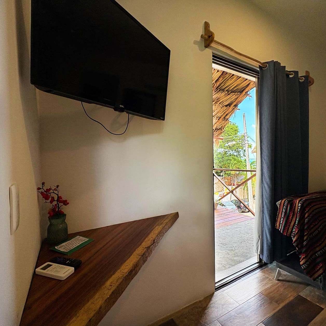 Une pièce dotée d’un téléviseur à écran plat fixé au mur, d’un banc en bois, d’une petite table avec un vase et une carte, et offrant une vue sur l’extérieur par une porte munie de rideaux.
