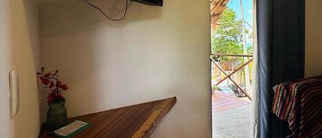 Une pièce dotée d’un téléviseur à écran plat fixé au mur, d’un banc en bois, d’une petite table avec un vase et une carte, et offrant une vue sur l’extérieur par une porte munie de rideaux.