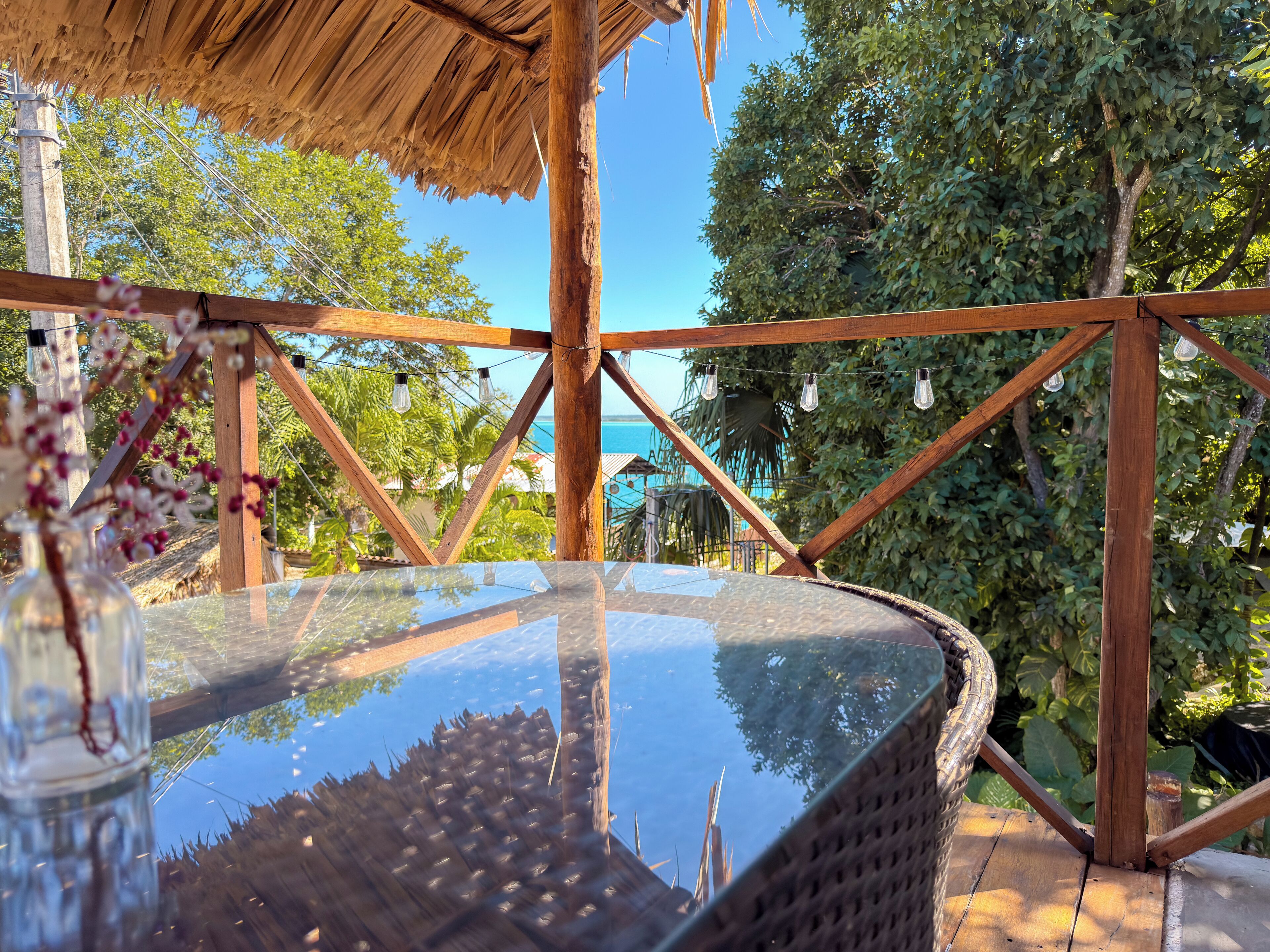 A glass-top table with a woven wicker frame, supported by wooden beams, under a thatched roof structure.