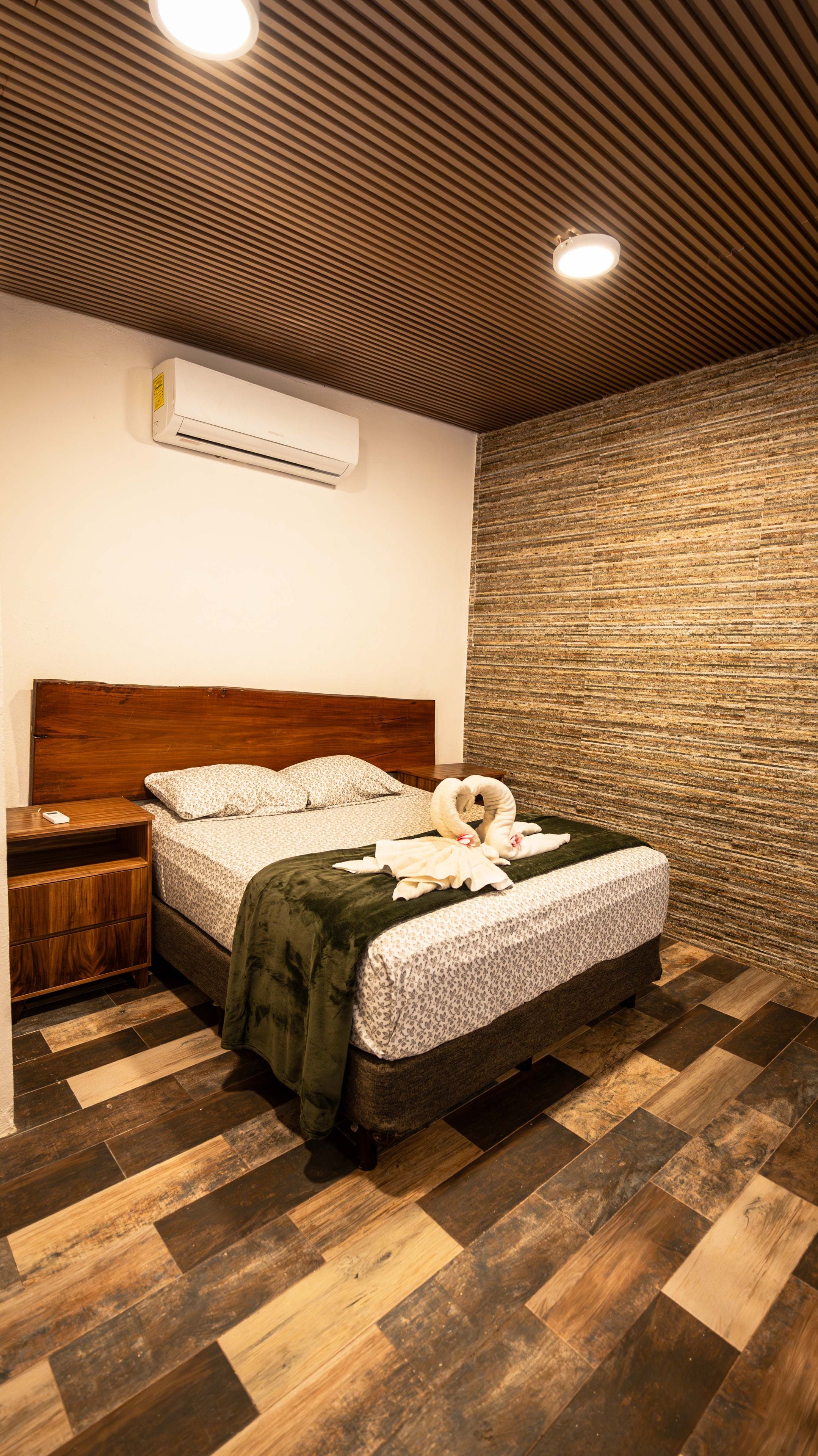 A hotel room with a wooden headboard, a bed with a floral bedspread, a green throw blanket, and a wall-mounted air conditioning unit.