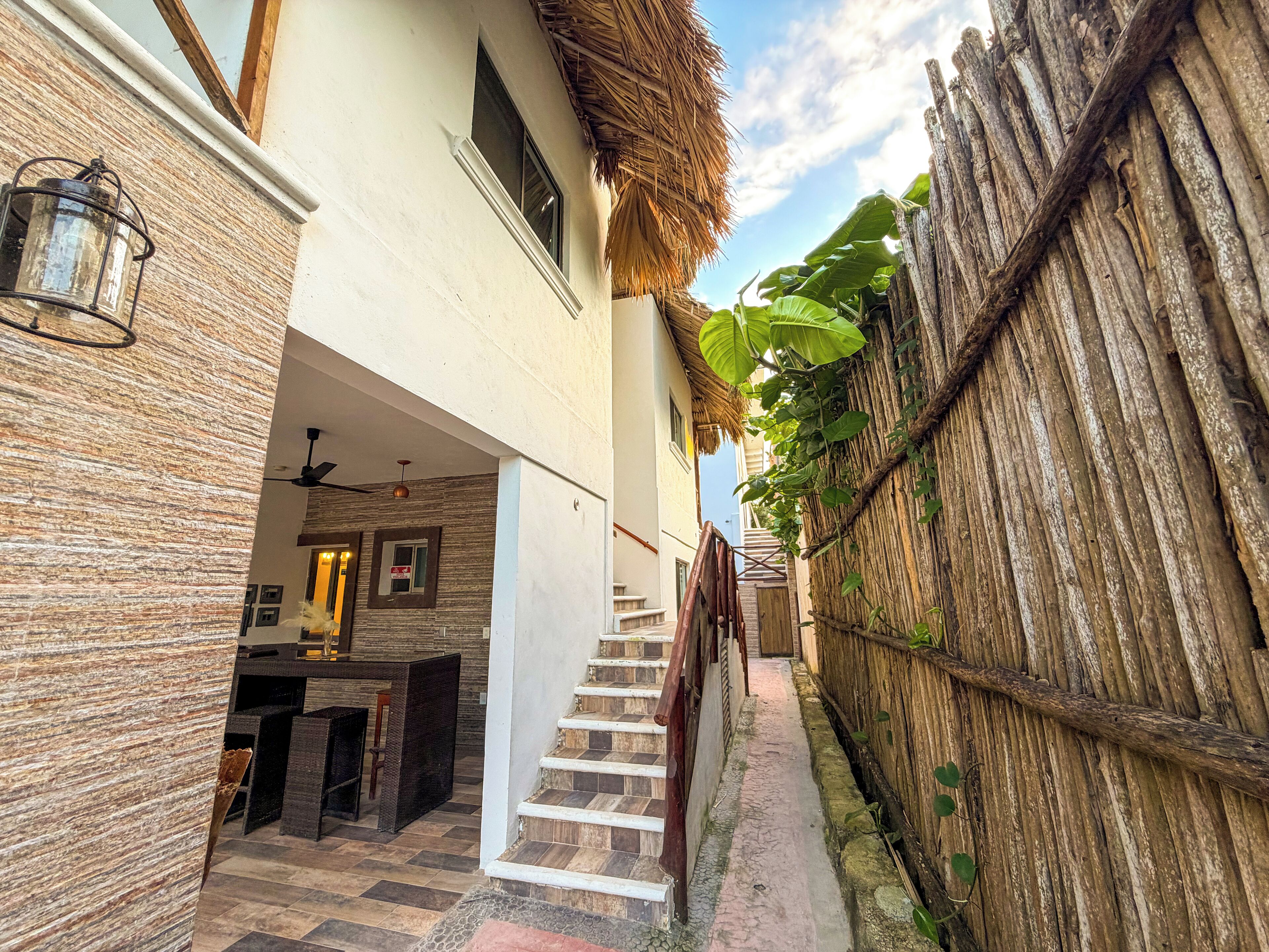 A narrow outdoor corridor with a staircase leading to a covered area, a wooden fence, and a thatched roof structure.