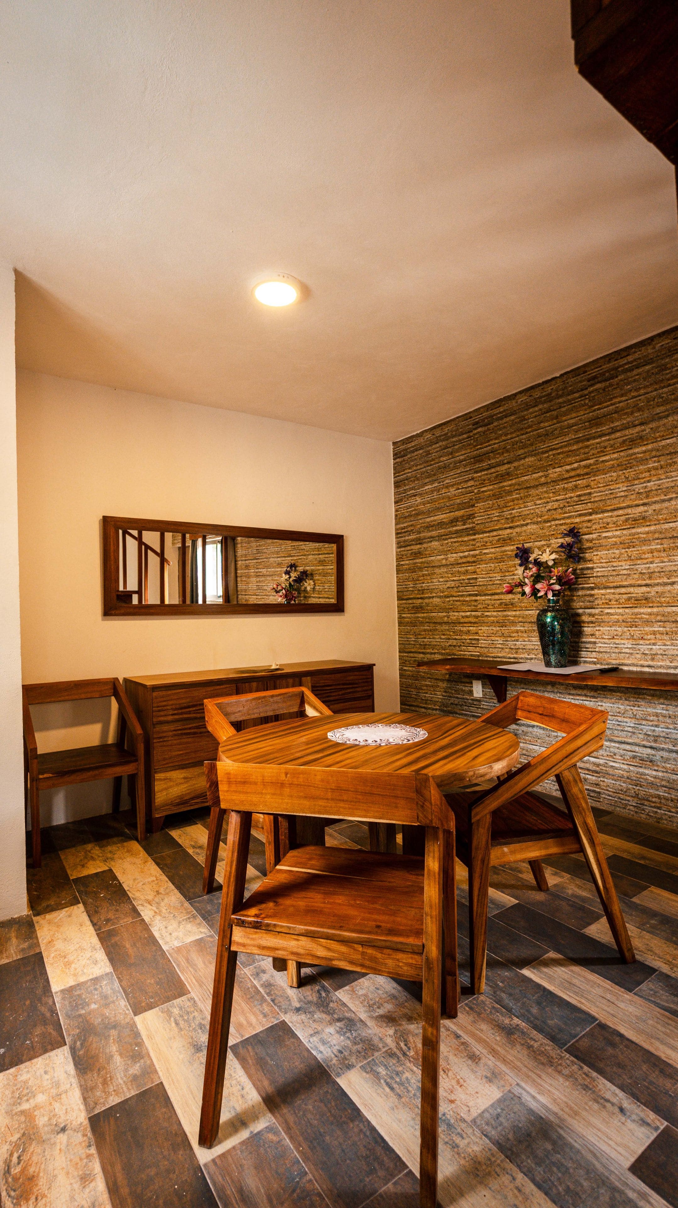A dining area with a wooden table and chairs, a bench, a mirror, and a wall-mounted vase with flowers.