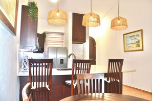A dining area with wooden chairs and a table, a kitchen with a refrigerator and cabinets, and hanging wicker lamps. - Oceanview Hideaway Apartment, Steps from the Beach (Cabarete)