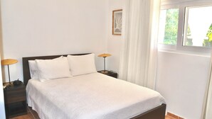 Una cama bien hecha con una colcha blanca, dos lámparas de noche y una ventana con cortinas.