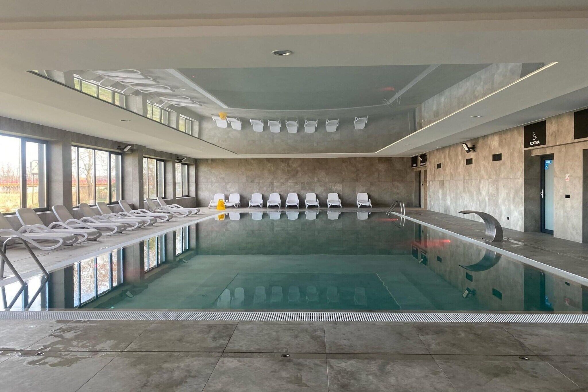 A large indoor swimming pool with multiple lanes, sun loungers along the sides, and a view of the outdoors through large windows.