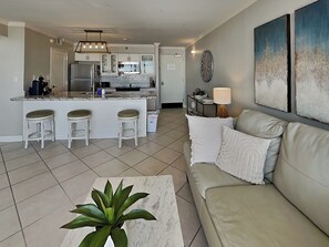 A modern kitchen with white cabinets, a central island with bar stools, and a dining area with a sofa and a lamp. - San Luis 736 - Oceanfront Paradise | Great Views (Galveston)