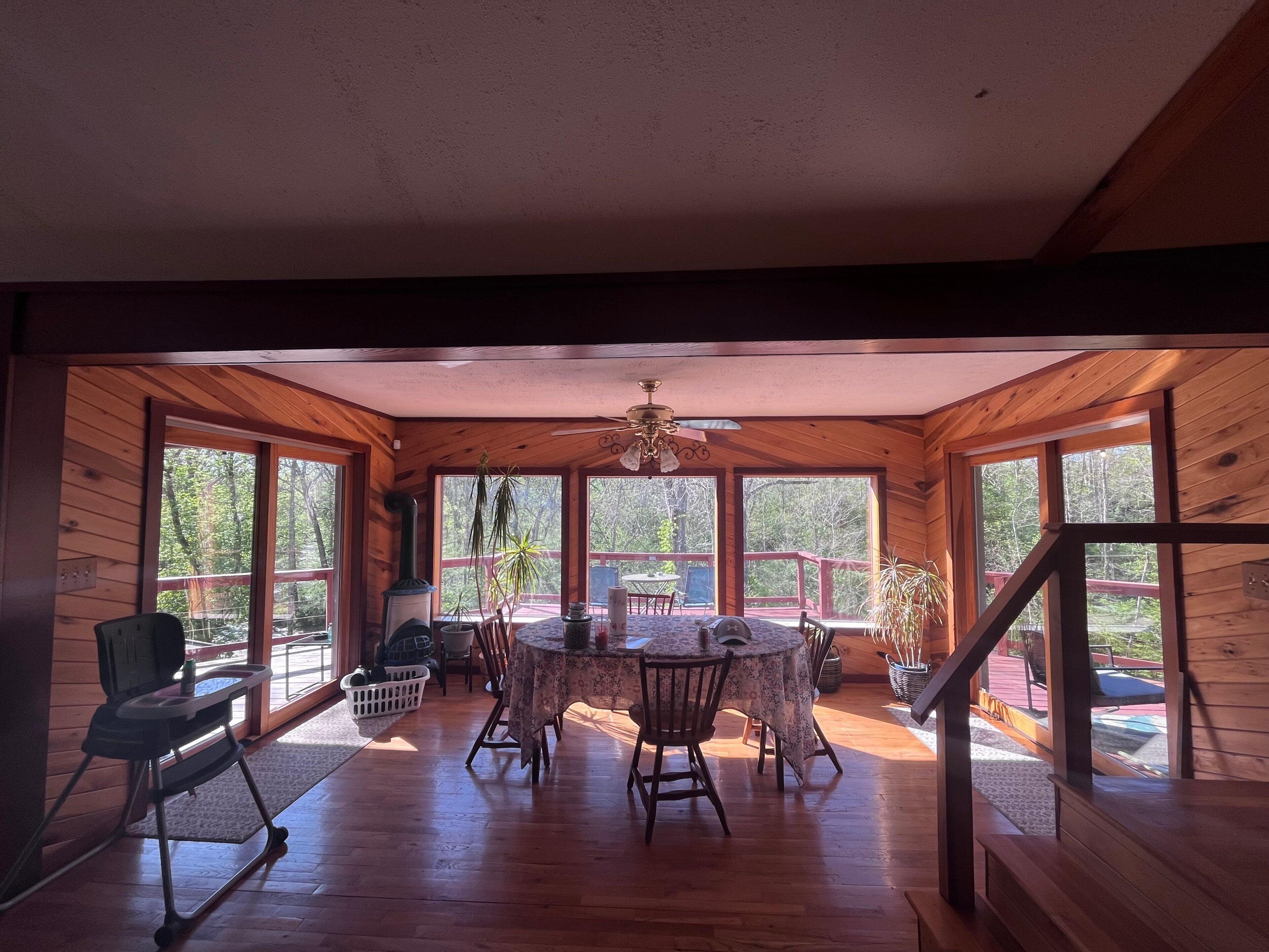 A wooden interior with a dining table and chairs, a ceiling fan, and large windows.