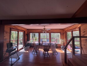 A wooden interior with a dining table and chairs, a ceiling fan, and large windows.