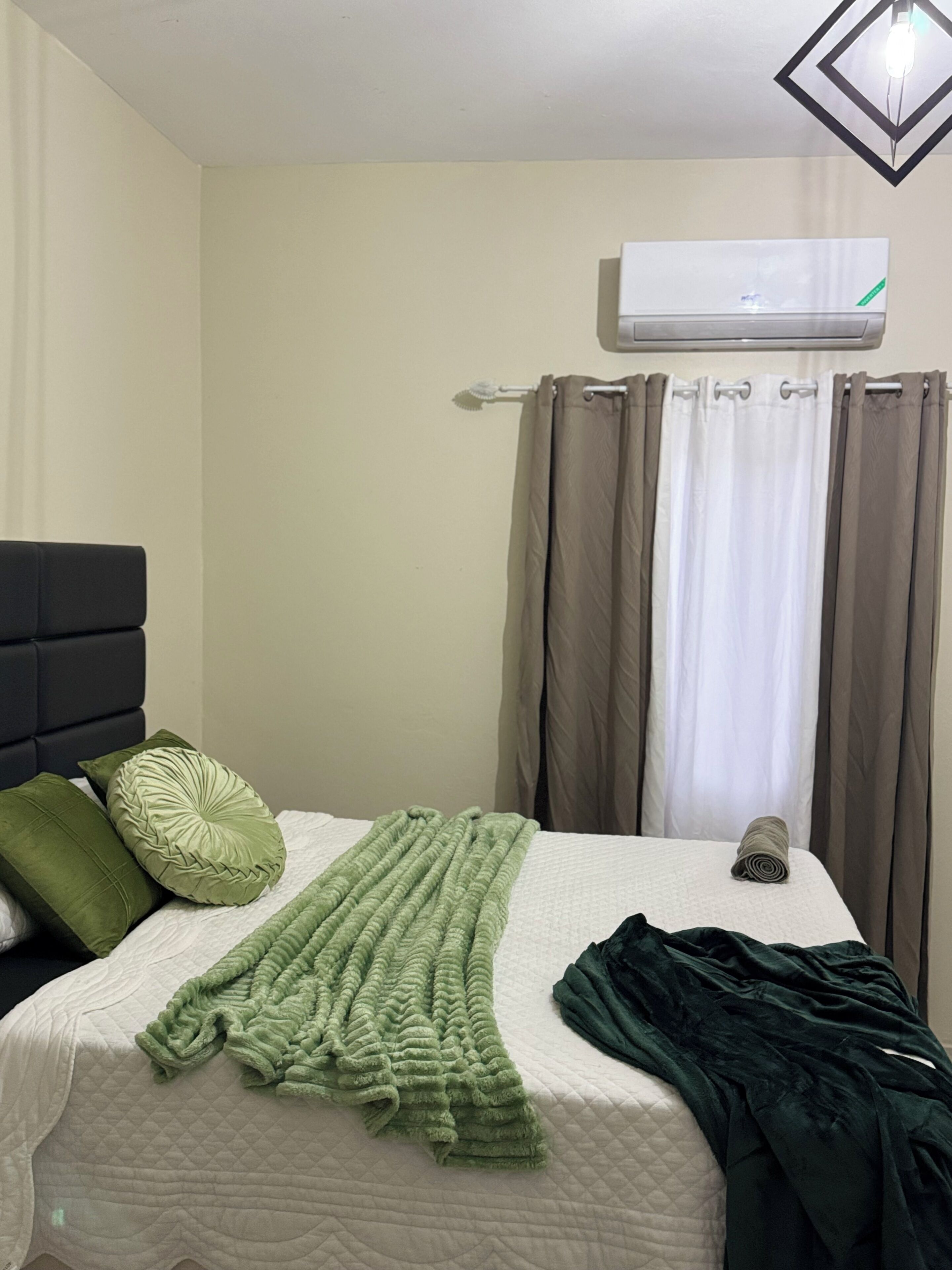 Une chambre à coucher comprenant un lit, une tête de lit, une couverture, des oreillers, une serviette enroulée et un climatiseur fixé au mur.