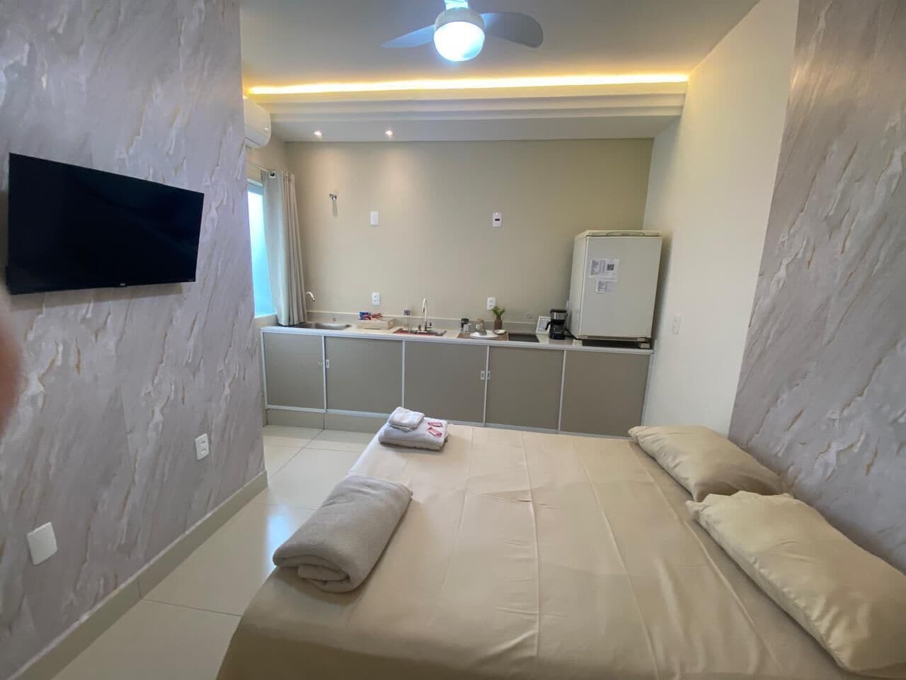 A hotel room with a bed, a flat-screen TV mounted on the wall, a kitchenette area, and a ceiling fan.