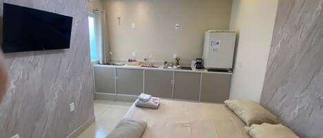 A hotel room with a bed, a flat-screen TV mounted on the wall, a kitchenette area, and a ceiling fan.