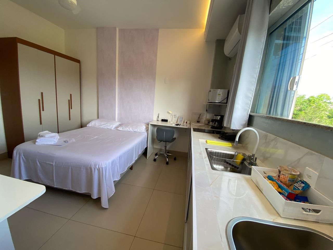 A hotel room with two beds, a desk, a chair, a kitchenette, and a window.