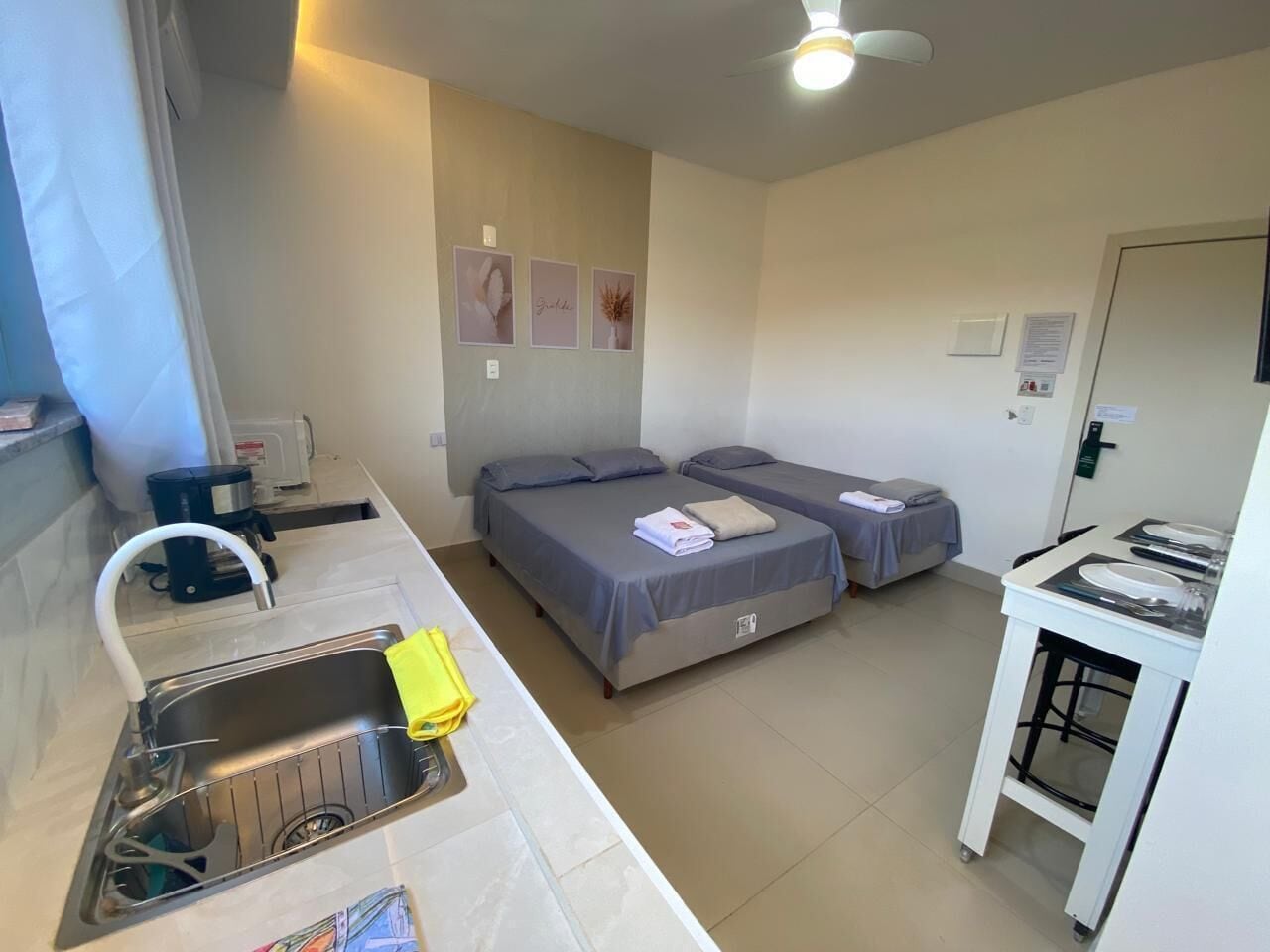 A compact hotel room with a kitchenette, two beds, a ceiling fan, and a small dining area.