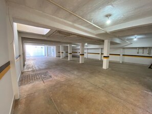 A spacious indoor parking area with concrete floors and white pillars, featuring a skylight and a visible exit. - Hotel Araujo's Suítes (Passos)