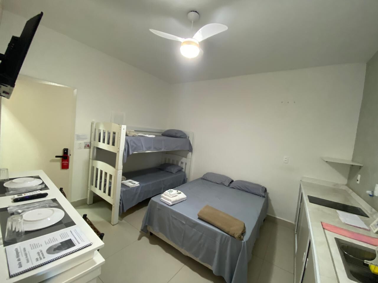Une chambre avec des lits superposés, un bureau avec une chaise, un ventilateur de plafond et une petite table avec une lampe.