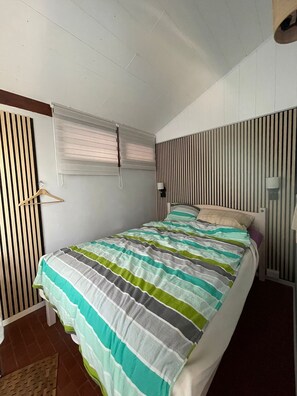 Ein Schlafzimmer mit Bett, Fenster mit Jalousien, Nachttisch und Wand mit vertikalen Linien.