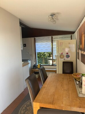 Ein Essbereich mit Holztisch, Stühlen, einem Kühlschrank und einem Fenster mit Blick ins Grüne.