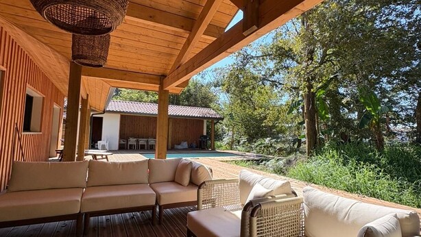 Una terraza de madera con mobiliario exterior, incluyendo un sofá y sillas, bajo un área techada con una lámpara de techo de fibras trenzadas.