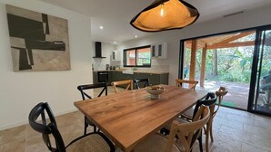 Un comedor moderno con una mesa y sillas de madera, una lámpara colgante y vistas a la vegetación a través de amplias puertas correderas.