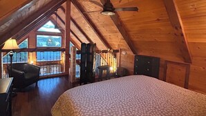 A bedroom with a wooden ceiling, a bed with a floral bedspread, a desk with a lamp, and a view of a snowy landscape through a window. - The Rustic Roost (Brigus Junction)