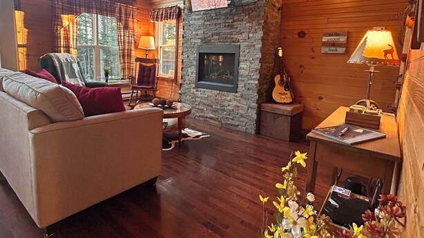 A cozy cabin interior with a stone fireplace, wooden walls, a sofa, and a dining table. - The Rustic Roost (Brigus Junction)