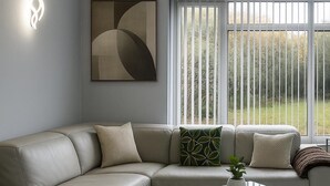 A modern living room with a sectional sofa, a glass coffee table, and a large window with blinds. - Charming 3BR Sleeps 7 Contractor Stay Driveway (Hale Barns)