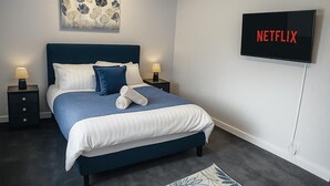 A modern bedroom with a bed, bedside tables, a wall-mounted TV, and a decorative painting. - Charming 3BR Sleeps 7 Contractor Stay Driveway (Hale Barns)