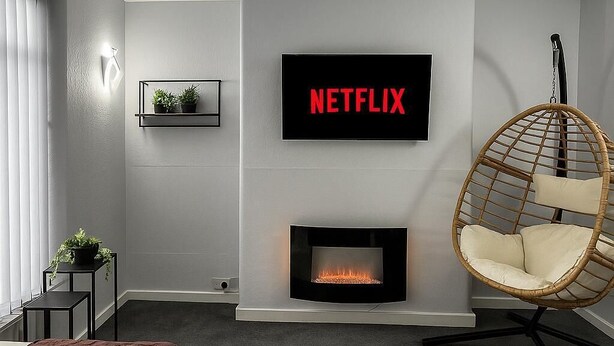 A modern living room with a fireplace, a mounted TV displaying 'NETFLIX', a hanging chair, and a glass coffee table with a vase and candles. - Charming 3BR Sleeps 7 Contractor Stay Driveway (Hale Barns)