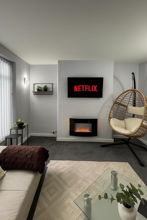 Una sala de estar moderna con chimenea, un televisor montado mostrando 'NETFLIX', una silla colgante y una mesa de centro de vidrio con un florero y velas.