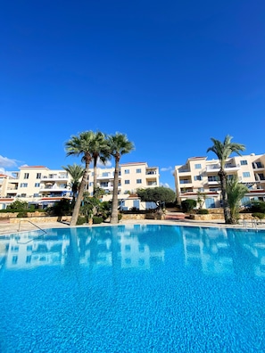 A swimming pool with clear blue water, surrounded by palm trees and a row of multi-story buildings with balconies. - 06 KPF - Stay Vera s Apartment (Paphos)