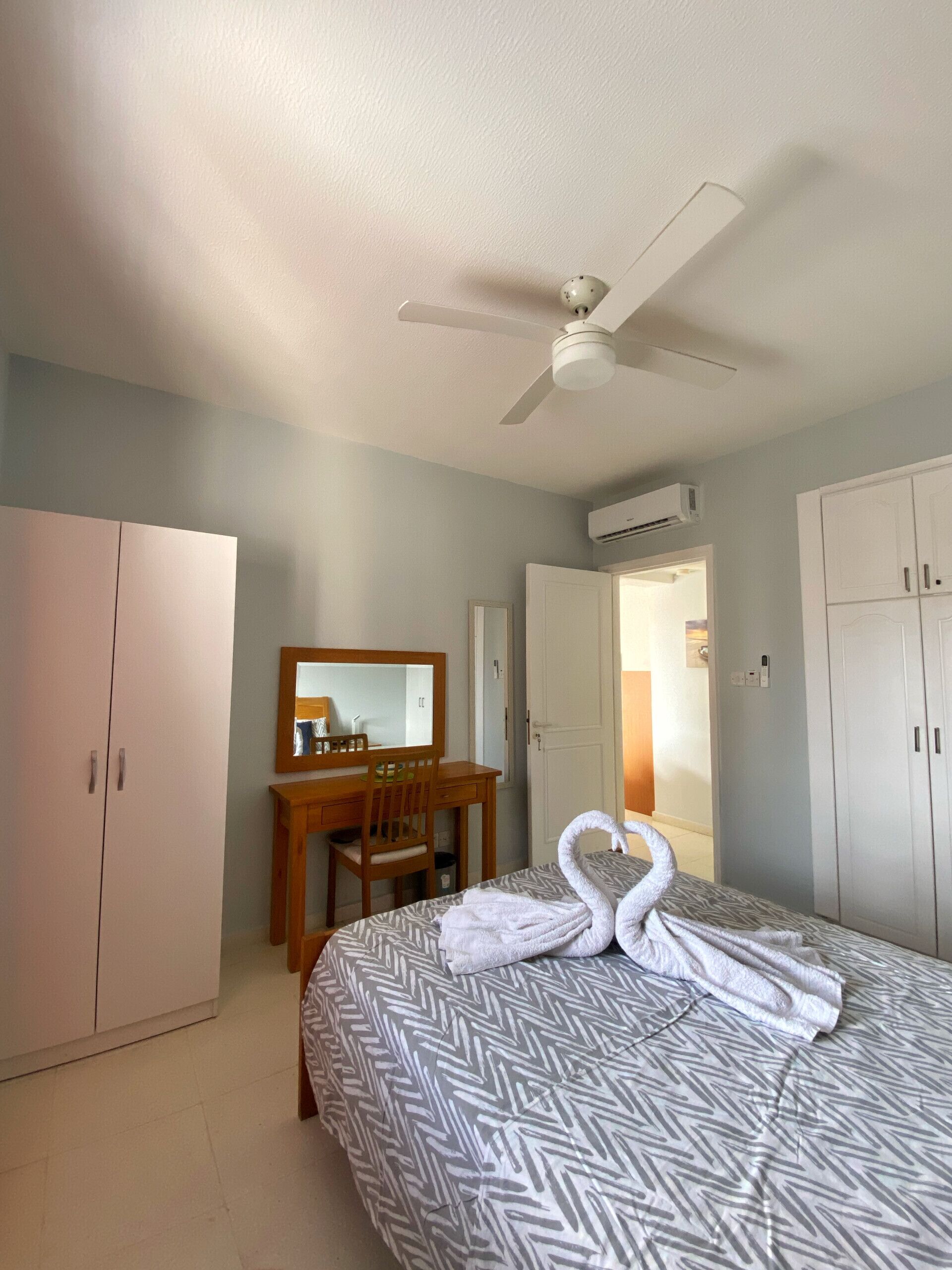A hotel room with a bed, a desk with a chair, a wardrobe, and a ceiling fan.