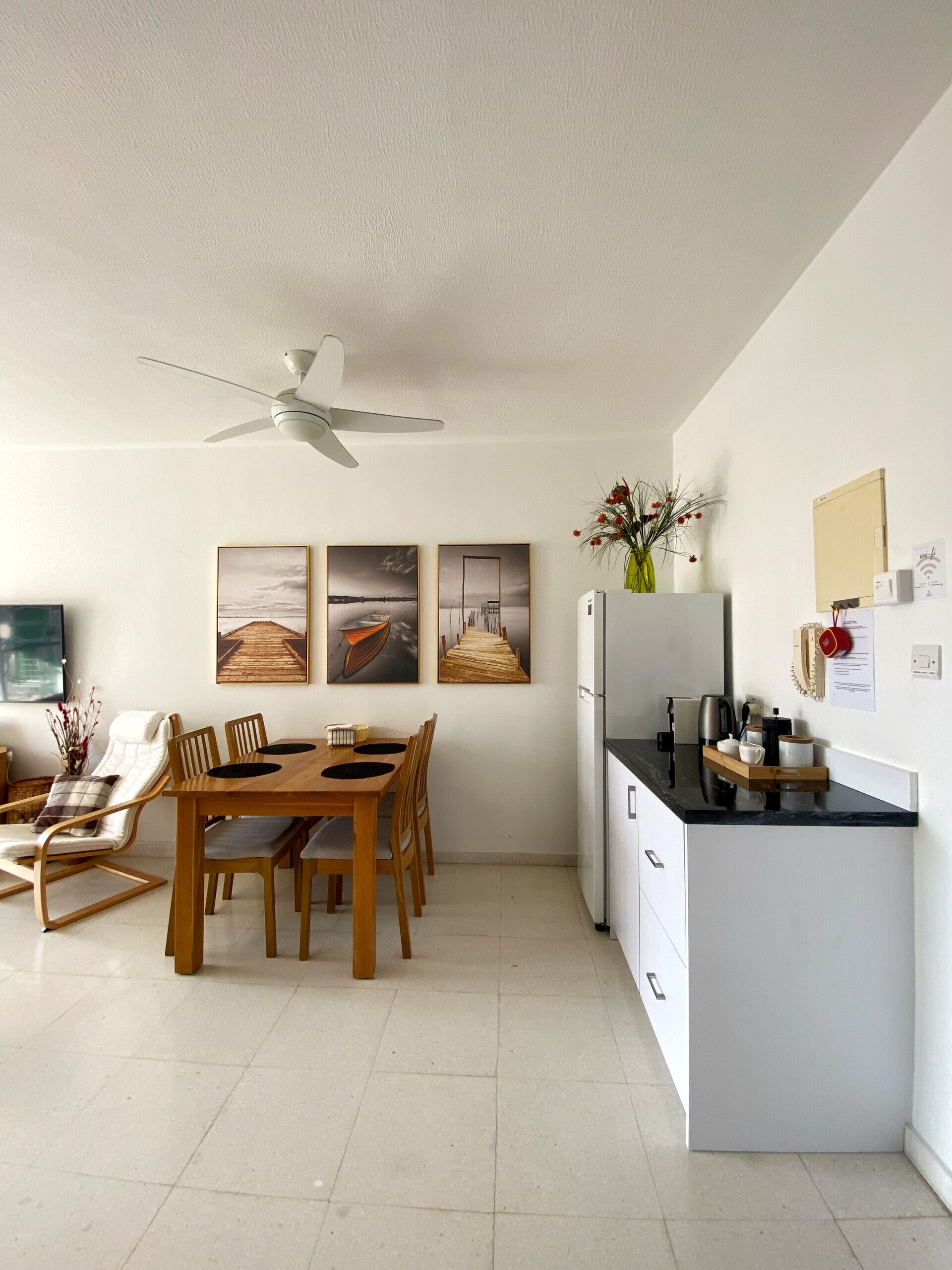 A modern kitchen with a white refrigerator, a coffee machine, and a wooden dining table with chairs. There are three framed pictures on the wall above the table.