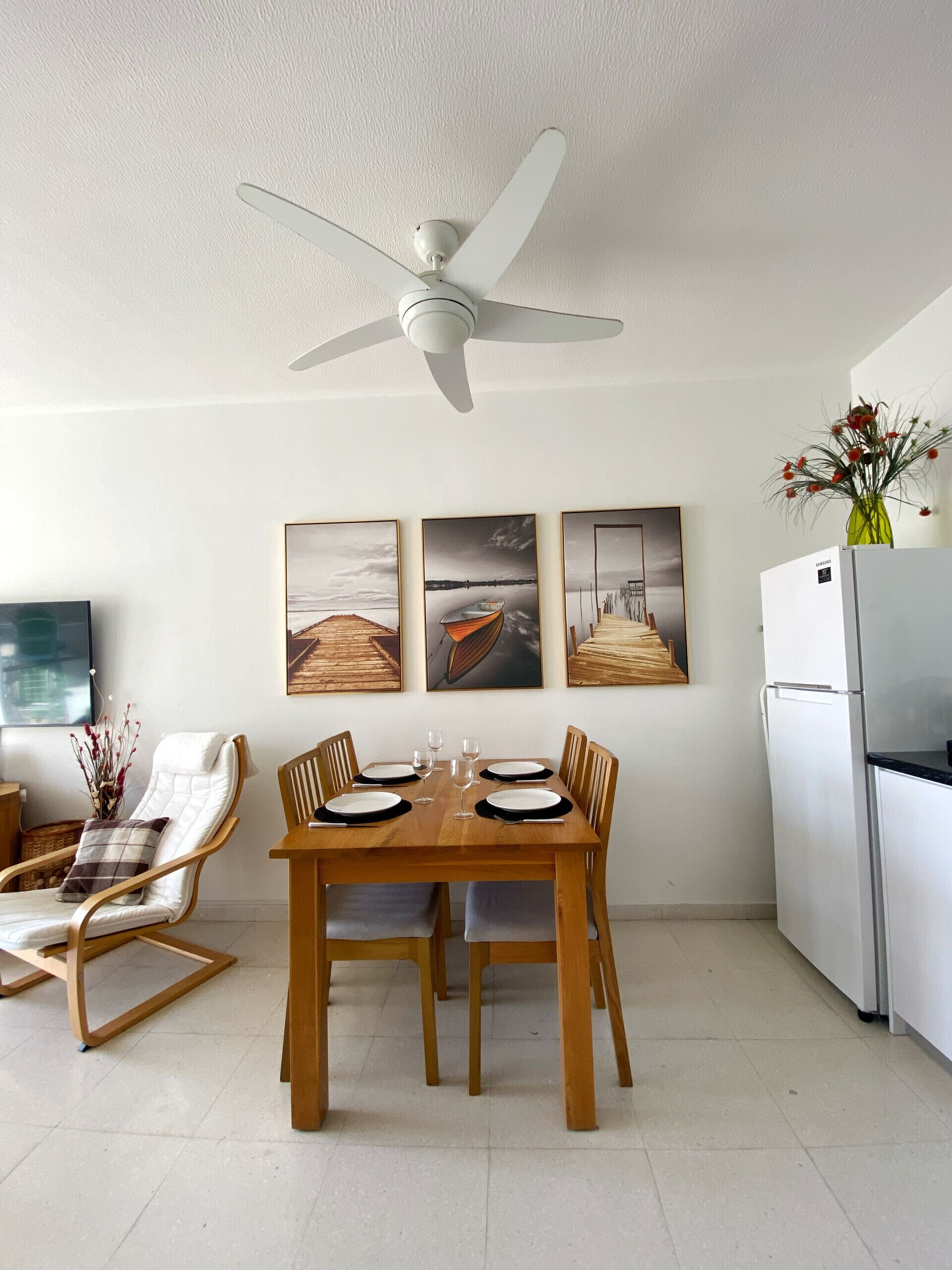 Un coin repas avec une table en bois dressée pour quatre personnes, un ventilateur de plafond et trois tableaux encadrés accrochés au mur.
