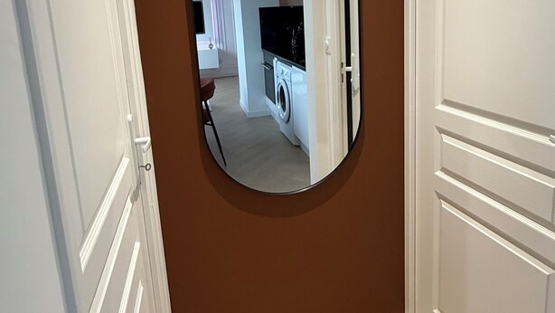 Un couloir avec un mur marron, un miroir rond et une porte donnant sur un espace cuisine.
